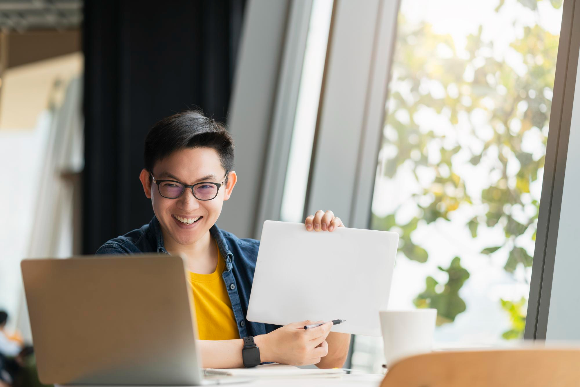 Asian male with glasses holding up a small whiteboard and pointing to it with a pen, while facing a laptop. (VitaCVs Contact Us Header)